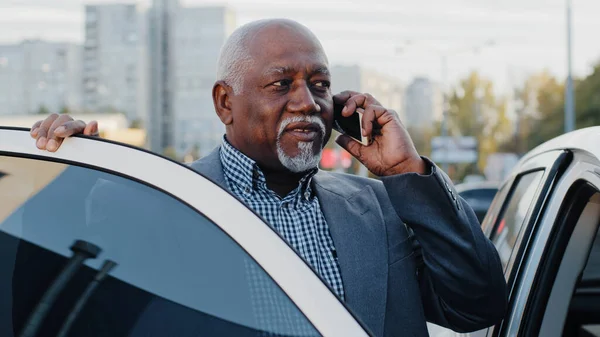 Mature african american businessman standing near car outdoors in parking lot speaks on mobile phone elderly friendly man communicates on telephone answers call using smartphone talking by cellphone — 图库照片