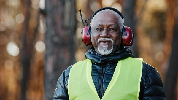 Elderly man professional forestry engineer in protective soundproof headphones standing in forest looking at camera mature african american foreman sawmill worker in uniform smiling posing outdoors —  Fotos de Stock