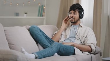 Young cheerful active hispanic guy having fun bearded arabian man lying on couch at home in apartment listening to music on headphones online singing song enjoying holiday radio audio sound indoors