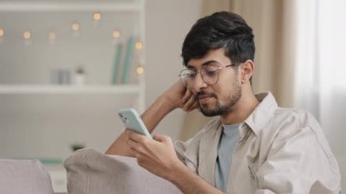 Yakın plan Arap kökenli genç adam gözlüklü erkek kullanıcı blogcusu cep telefonuyla evde oturuyor internette sohbet ediyor internetten mesaj yazıyor gülümsüyor akıllı telefondan ücretsiz wi-fi kullanıyor.