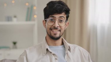 Male portrait close-up bearded face millennial arabic indian man guy with glasses looking at camera smiling waving nods head answering yes positive decision agreement support approval sitting at home