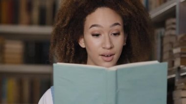 Close-up african american girl student hiding behind textbook reading book emotional young woman stands in library attentively reads novel surprised shocked by unexpected ending feeling bewildered