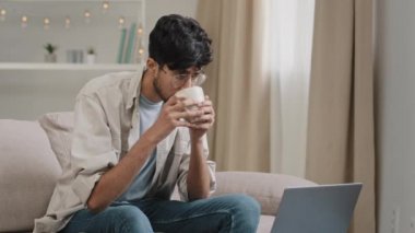 Young freelancer male business man bearded guy in glasses drinks tea coffee delicious drinking at home sitting on couch watching movie in laptop online video reads news on network receives message