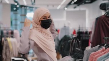 Kaygısız genç Arap kadın alıcı tıbbi koruyucu maske takıyor alışveriş merkezindeki giyim mağazasında alışveriş yapıyor tesettürlü mutlu Müslüman kız dans ediyor hareketli dans ediyor indirimlerden keyif alıyor