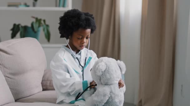 Serious african american kid girl sitting in room on couch in medical uniform pretending to be doctor little child playing veterinarian heals toy patient holding stethoscope listening sick teddy bear — Vídeos de Stock