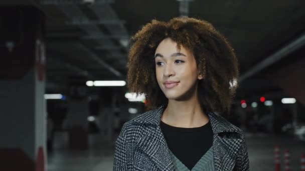 Όμορφη νεαρή σγουρά μαλλιά αφροαμερικανίδα κοπέλα χιλιετή επιτυχημένη κομψή γυναίκα γυναίκα που περπατά στο πάρκινγκ του εμπορικού κέντρου επιχειρήσεων κοιτάζοντας κάμερα κινείται μέσα σε εσωτερικούς χώρους — Αρχείο Βίντεο