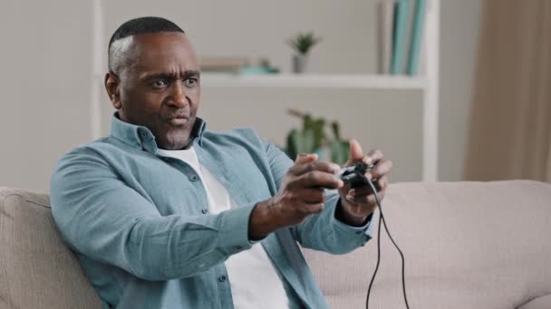Зрелый африканский американец сидит на диване в комнате внимательно играет в видеоигры на консоли взрослый эмоциональный мужчина играть в игры использует джойстик контроллера управления сосредоточены на конкуренции наслаждается досугом — стоковое видео