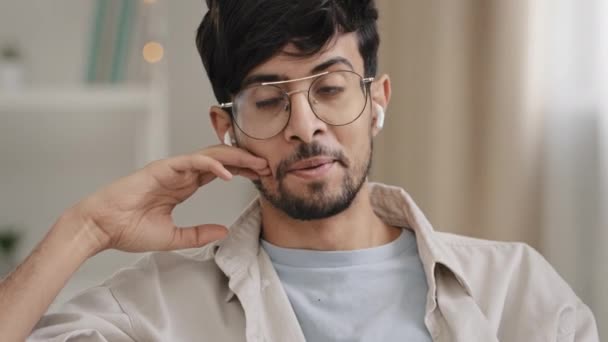 メガネを持つクローズアップアラビア語の若いひげを生やした男は、ワイヤレス現代のヘッドフォンを使用してオンライン通話を話します。肖像画ヒスパニック男回答コールに自宅でチャットで友人とリモートで笑顔会話 — ストック動画