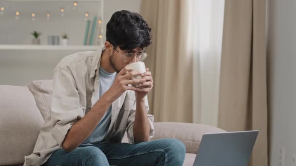 Joven freelancer hombre de negocios hombre barbudo en gafas bebidas té café delicioso beber en casa sentado en el sofá viendo la película en el ordenador portátil video en línea lee noticias en la red recibe el mensaje — Vídeo de stock