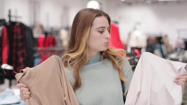 Young stylish beautiful girl woman female client buyer customer stands in clothing store shop makes choice choosing between two trendy sweaters jumpers thinking doubts shopping discounts sales concept — Stok video