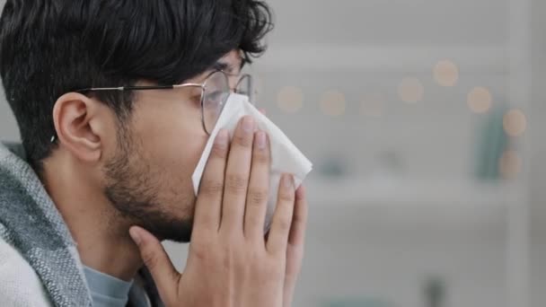 Primer plano cara masculina vista lateral enfermo árabe hispano hombre barbudo con gafas envueltas en manta sufre de secreción nasal estornudos alergia toallitas mocos con papel servilleta enfermedad respiratoria infección — Vídeo de stock