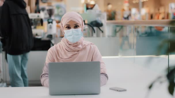 Young muslim businesswoman in hijab in medical mask sits in public place working on laptop arab girl student studying online communicates in computer chat uses new app shows ok sign gesture approval — Stockvideo