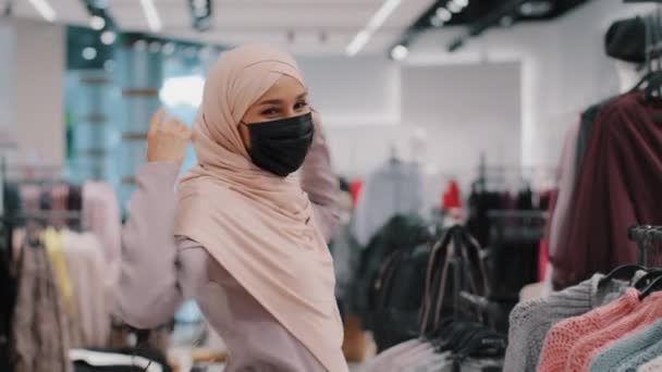 Carefree young arab woman buyer wearing medical protective mask walking shopping on clothing store in mall happy muslim girl in hijab whirls dancing actively moving rejoices sale enjoying discounts — Stockvideo