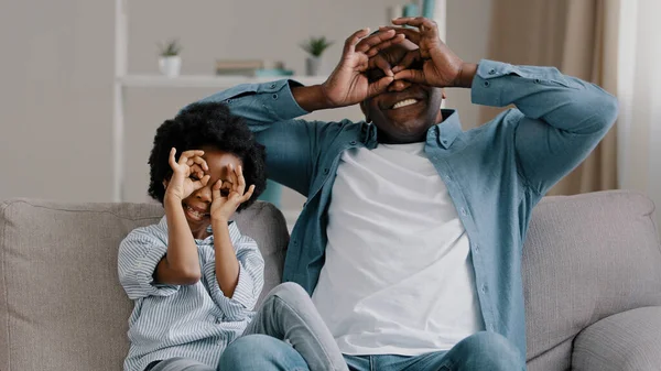 Šťastný veselý africký americký starší otec hrát s roztomilou dcerou sedí v pokoji na pohovce usmívající se táta a holčička dělat s rukama legrační brýle masky předstírat, že nosí brýle oklamání — Stock fotografie