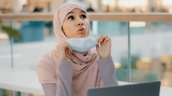 Giovane araba donna d'affari in hijab lavora al computer portatile si guarda intorno ragazza musulmana abbassa espira maschera medica con sollievo divertente graffi naso mette respiratore sui volti donna stanca di restrizioni di quarantena — Foto Stock