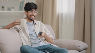 Arab man bearded guy with glasses resting at home sitting on couch drinking tea coffee watching funny movie championship sports game TV rejoices at victory of favorite team laughing fun sincerely