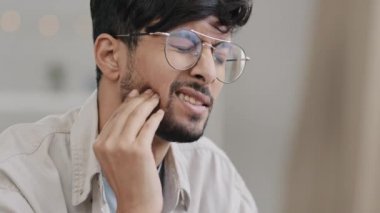 Close-up male sad face hispanic arab bearded man with glasses suffering guy feels toothache discomfort in gum caries problems orthodontist help need holding to cheek feeling unwell illness pain ache