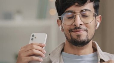 Close-up arabic spaniard man bearded guy in glasses wears wireless modern headphones listening to music with phone dancing enjoying high quality audio sound mobile application website with songs