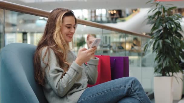 Веселая счастливая молодая девушка покупатель шопоголик женщина сидит в торговом центре с пакетами глядя на телефон побеждает радуется скидки успешный заказ выбирает одежду онлайн победу да жест — стоковое видео