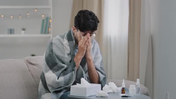 Arab bearded sick sad man with glasses sits on couch at home suffers from runny nose flu disease coronavirus pandemic covid epidemic sneezes. Unwell guy feeling bad fever virus illness symptoms indoor — Stok video