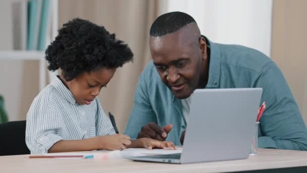 African American family kid girl sitting in room at desk doing homework father approaches and points out mistake child laughs gives pen teacher dad helps daughter with homeschooling during quarantine — Stok video