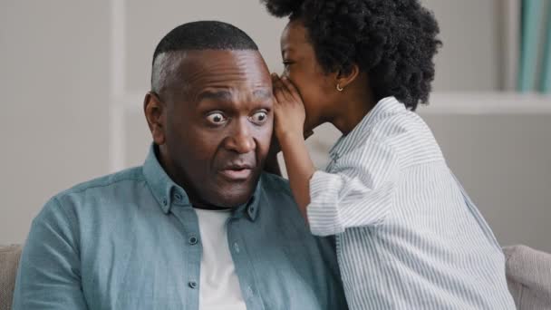 Little African American girl whispers in ear reveals secret to daddy daughter shares secrecy gossip confidential information mature father sitting on couch surprised from conversation opens eyes wide — Vídeo de stock