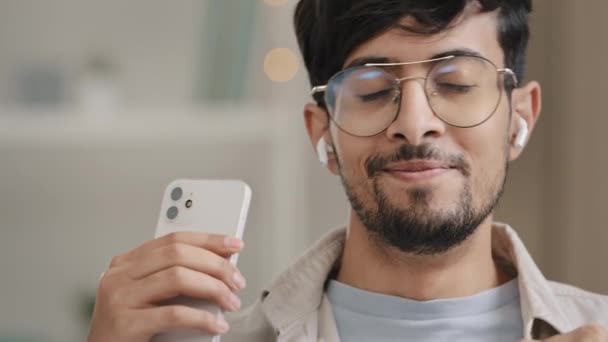 Крупним планом арабська іспанська людина бородатий хлопець в окулярах носить бездротові сучасні навушники, слухаючи музику з телефонними танцями, насолоджуючись високоякісним аудіо звуковим мобільним додатком веб-сайтом з піснями — стокове відео