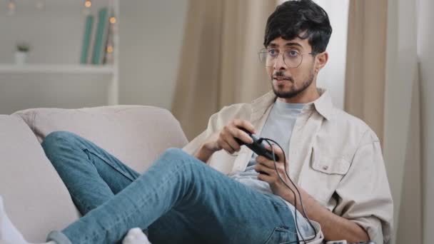 Arab guy bearded man with glasses lying on couch at home playing console online video game competition emotionally winning yes hand gesture victory having fun with controller addiction to videogaming — Stockvideo