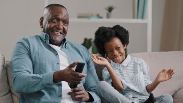 Close-up šťastný africký americký otec a dcera se směje sledování zábavné televizní show film relaxační na gauči muž drží dálkové ovládání ukazující palec nahoru gesto souhlasu táta a holka baví — Stock video