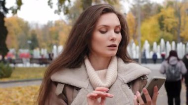 Pivoting portrait of discompose brunette American woman holding smartphone in autumn park centre and looking away. The girl in pullover is looking out for someone on street with cell in his hands.