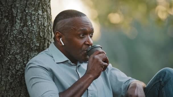 Zdjęcie głowy Starszy Afroamerykanin czarny biznesmen siedzący pod drzewem, odpoczywający w parku miejskim podczas przerwy. Dorosła, uśmiechnięta osoba nosząca słuchawki bezprzewodowe pije kawę lub herbatę. — Wideo stockowe
