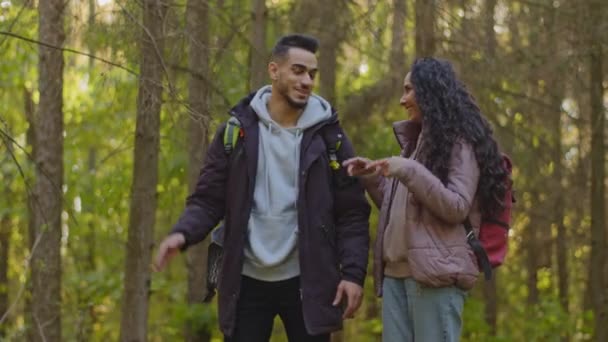 Urban people got lost in wood. Funny cheerful Multinational couple of diverse friends waving hands, showing correct route, laughing. Hispanic woman and arabic man walking having fun in autumn forest — Stock video