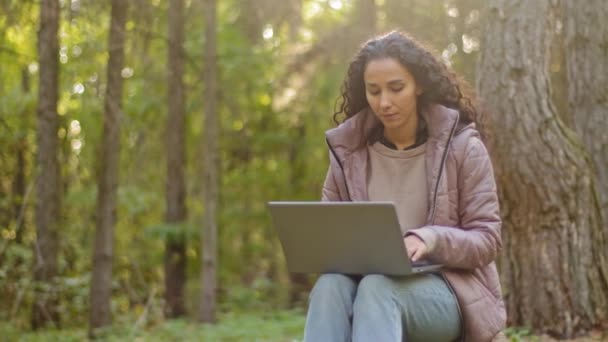 Mooie jonge Spaanse vrouw met lang krullend haar buiten in het najaar bos met behulp van laptop. Millennial vrouwelijke reizen blogger typen op zoek gepensioneerd naar computer scherm op bomen achtergrond — Stockvideo