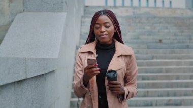 Frustrated African American businesswoman walking stairs with disposable cup of coffee and telephone. Sad young woman receiving unpleasant cancellation message, annoyed worried by bad news, problems