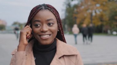 Closeup headshot happy attractive African American young woman standing outdoor in autumn holding smartphone, charming smiling millennial female student chatting on phone, having pleasant conversation