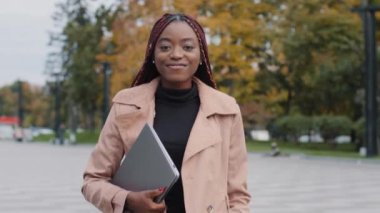 Beautiful smiling millennial African American female manager with laptop, successful businesswoman or student walking downtown in casual stylish autumn clothing. Happy young lady looking at camera 