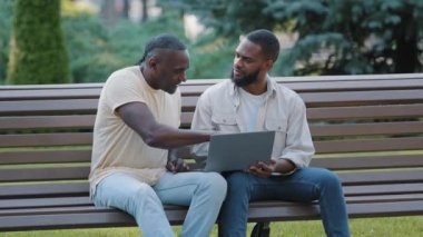 African American black male friends sitting in park looking at computer screen. Two men of different ages freelancer and customer argue laughing using laptop, discussing results of deal, good news