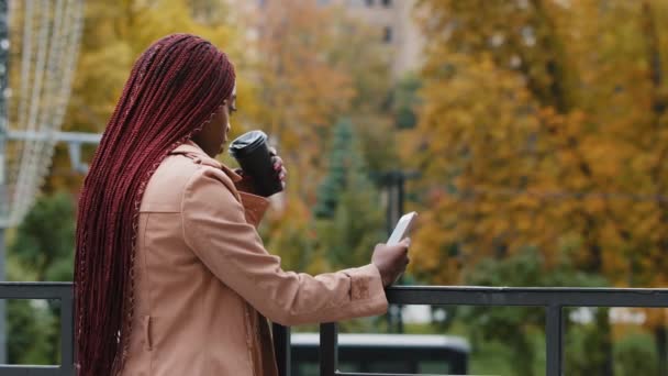 Young african american girl standing with phone and coffee on autumn day outdoors. Student waiting for meeting, female office employee resting during break, typing SMS on smartphone, checking mail — Stock video
