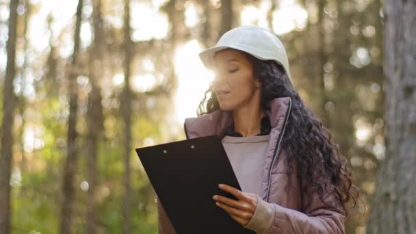 Millennial experienced Female technician with clipboard taking measures for reforestation of woodlands. Young indian Forestry engineer in hardhat in park. Supervising wildlife sanctuary checking trees — Vídeo de stock