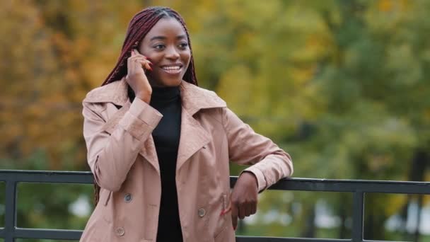 Alegre joven afroamericana mujer de pie al aire libre en el día de otoño felizmente charlando por teléfono celular. Chica estudiante sonriente, empleado de la oficina femenina toma llamada telefónica tiene una conversación agradable — Vídeo de stock
