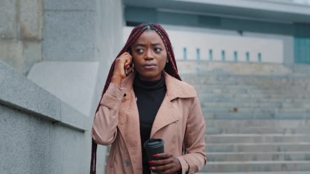 Anxious concerned African American young woman walking stairs with disposable cup of coffee and smartphone. Frustrated sad businesswoman has unpleasant cell phone conversation worried by bad news — Vídeo de Stock