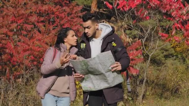 Multiracial couple millennial students walking in autumn forest young Indian man and hispanic woman traveling with backpacks holding using paper map looking for way make up route, tourist concept — Stock video