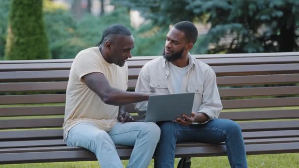 African American black male friends sitting in park looking at computer screen. Two men of different ages freelancer and customer argue laughing using laptop, discussing results of deal, good news — Vídeo de Stock