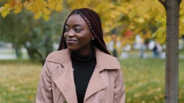 Female employee having break outdoor. Smiling happy dreamy African American young woman walking in city park in afternoon alone. Millennial student girl enjoying nature outing on cloudy autumn day