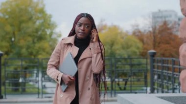 Successful attractive young african american lady, holding smartphone and document folder, assistant director, secretary, businesswoman, manager talking on phone outdoors, answering business call 