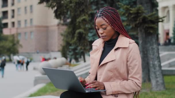 Focused attractive young african american lady businesswoman working on laptop sitting outdoors, female entrepreneur professional freelancer using computer software communicates in social networks — стоковое видео