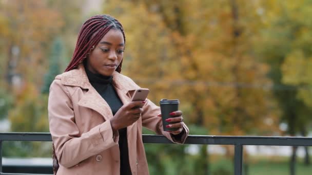 Beautiful young African American business woman office worker having break drinking coffee outdoor on autumn day. Long haired millennial girl looking at smartphone screen reading sms, checking mail — Stock video