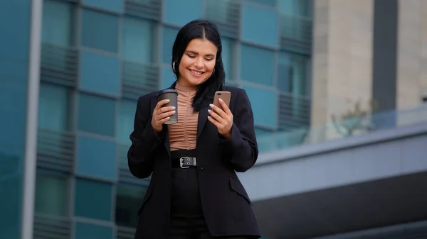 Joven mujer de negocios pasa tiempo libre en las redes sociales de Internet utilizando el teléfono inteligente utiliza servicios virtuales de compras en línea chat de texto de correo electrónico en teléfono chica hispana bebe café de la taza de papel al aire libre — Foto de Stock