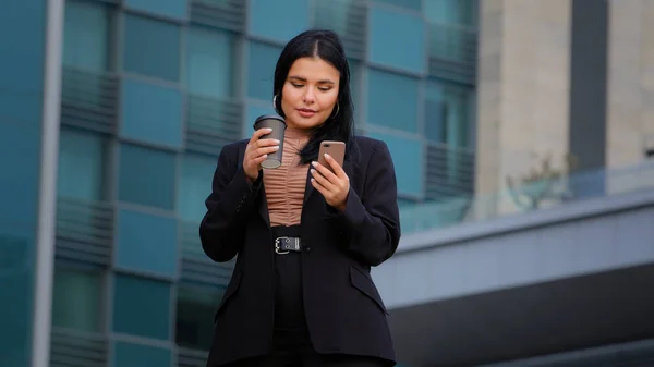 Joven mujer de negocios pasa tiempo libre en las redes sociales de Internet utilizando el teléfono inteligente utiliza servicios virtuales de compras en línea chat de texto de correo electrónico en teléfono chica hispana bebe café de la taza de papel al aire libre — Foto de Stock