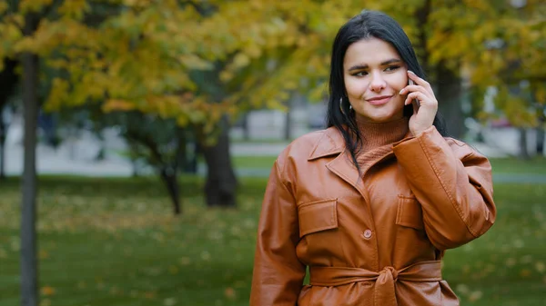 Young smiling woman outdoors in autumn park talking on mobile phone hispanic girl enjoys pleasant conversation communicates by telephone female customer makes order in online store using smartphone — Foto de Stock